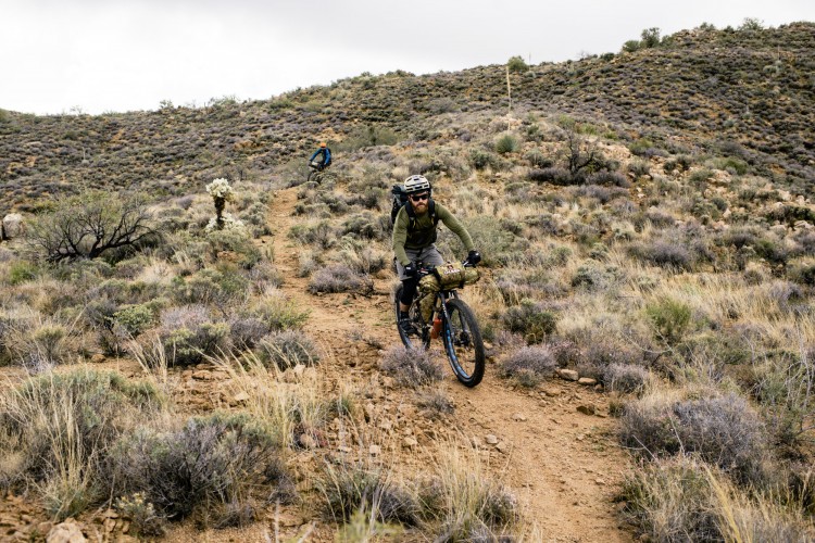 A person riding a mountain bike along a narrow dirt trail in a hilly, desert landscape, surrounded by sparse vegetation and shrubs. The sky is overcast, suggesting a cool and perhaps breezy day. Another cyclist is visible in the background, also navigating the trail.