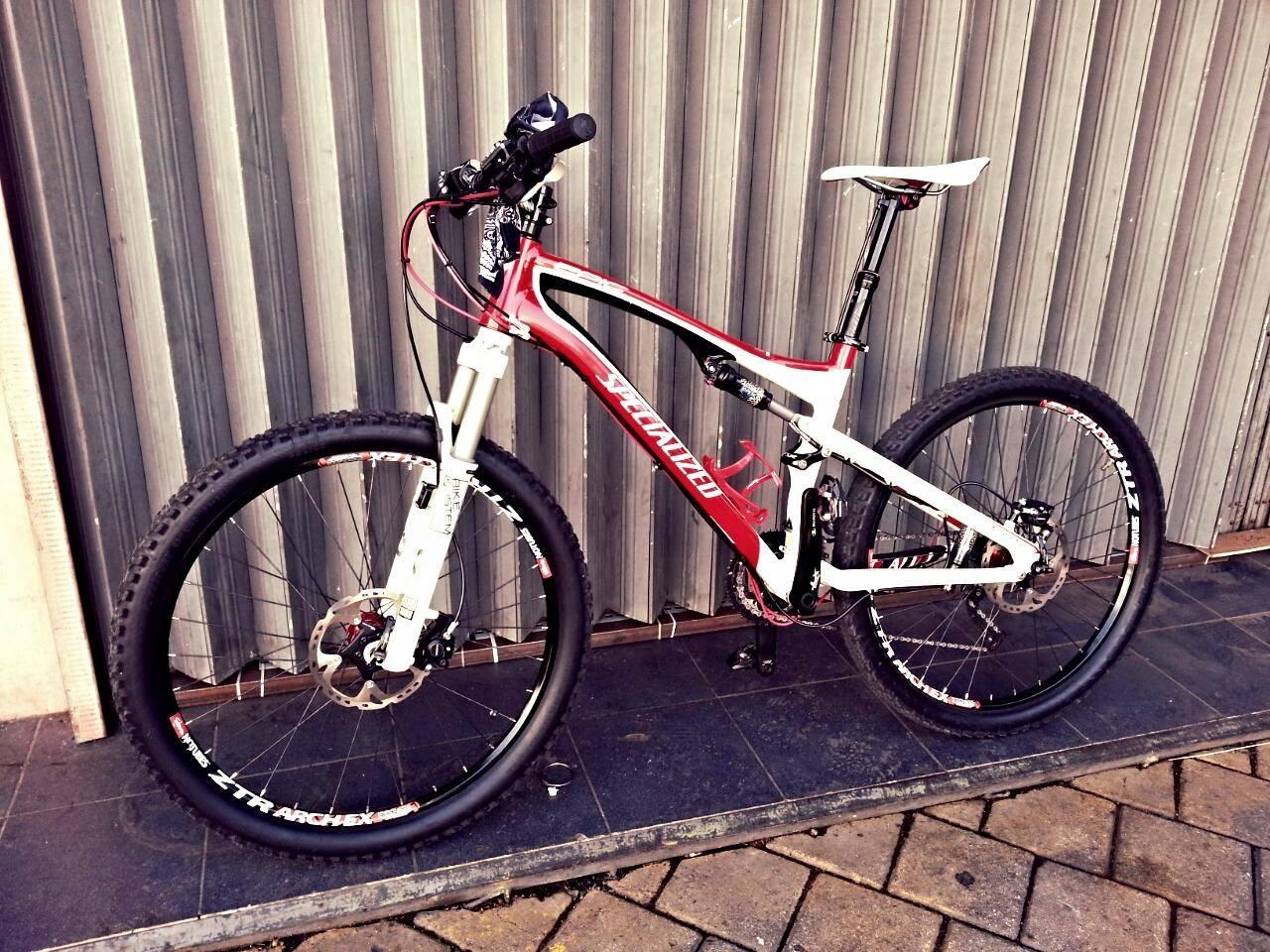 Specialized Epic Comp: A red and white mountain bike leaning against a corrugated metal wall, featuring a front suspension and large knobby tires. The bike is equipped with disc brakes and a sleek design, showcasing its sporty features. The ground is paved with square tiles.