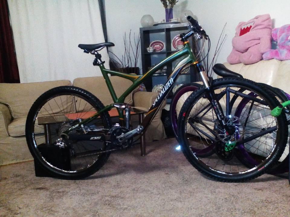 Specialized Stumpjumper: A specialized mountain bike with a green and black frame, positioned in a living room setting. The bike is leaning against a beige sofa, with a carpeted floor and various home decor items in the background, including a shelf with decorative objects and a plush toy. The image captures the bike's details, including its tires and components.