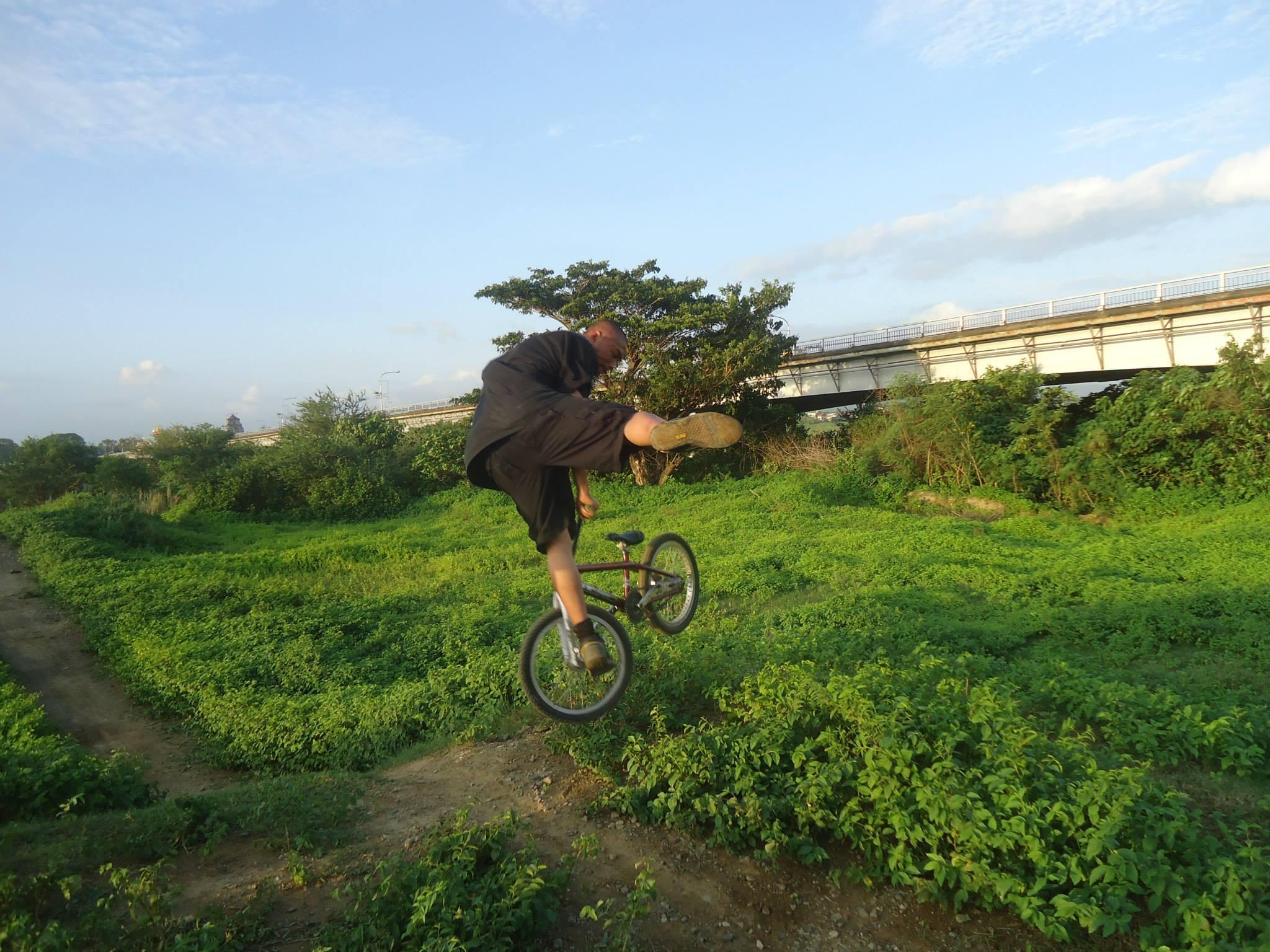 Specialized Stumpjumper: A person performing a BMX stunt in a lush green environment, with one foot raised and the bike's back wheel off the ground. In the background, a bridge can be seen crossing over a river, and the sky is partly cloudy.