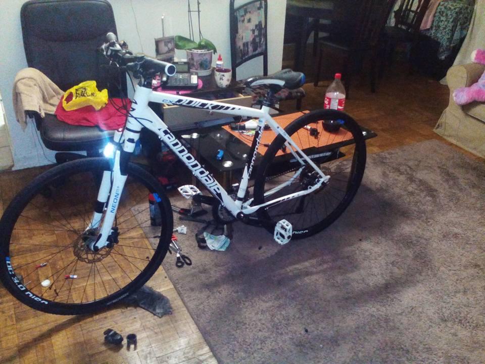 Specialized Stumpjumper: A white Cannondale bicycle is parked in a living room, surrounded by various household items. The bike features visible components such as a front light and distinctive black tires. In the background, there is a dining table, a chair, and scattered clothes, creating a casual and lived-in atmosphere.