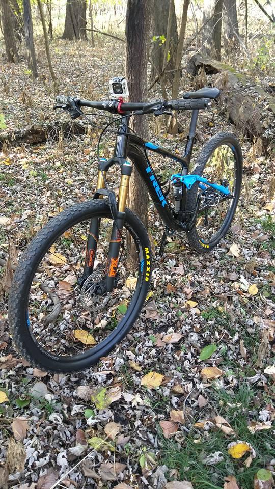 Trek Fuel EX 7 29: A mountain bike with a black frame and blue accents is leaning against a tree in a wooded area. The ground is covered with fallen leaves in various shades of brown and green, indicating autumn. A small action camera is mounted on the handlebars, suggesting the bike is ready for an outdoor adventure or recording a ride.