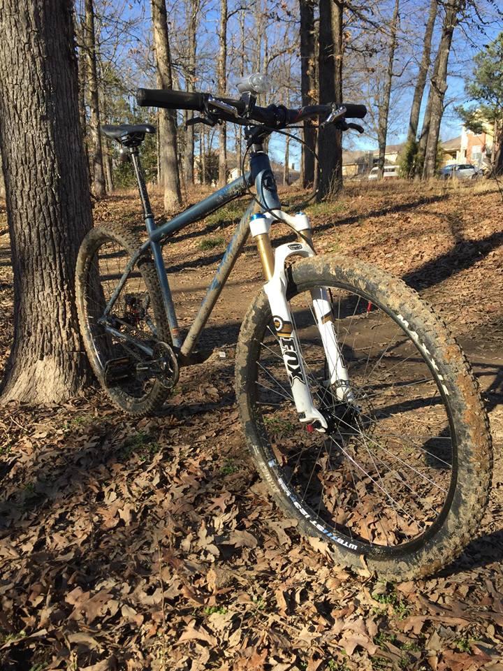 Kona Unit 29er: A mountain bike leaning against a tree, surrounded by fallen leaves. The bike features a lightweight frame, thick tires with mud on them, and a front suspension fork. In the background, a clear blue sky and bare trees suggest a natural outdoor setting.