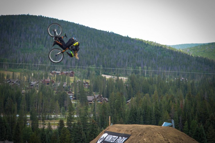 A mountain biker performs a trick in mid-air during a jump over a dirt ramp, with a forested mountain landscape and a few houses visible in the background.