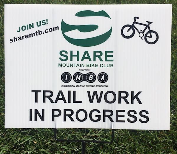 Sign indicating trail work in progress by the SHARE Mountain Bike Club, with a green logo and illustration of a bicycle, along with a website link for joining.
