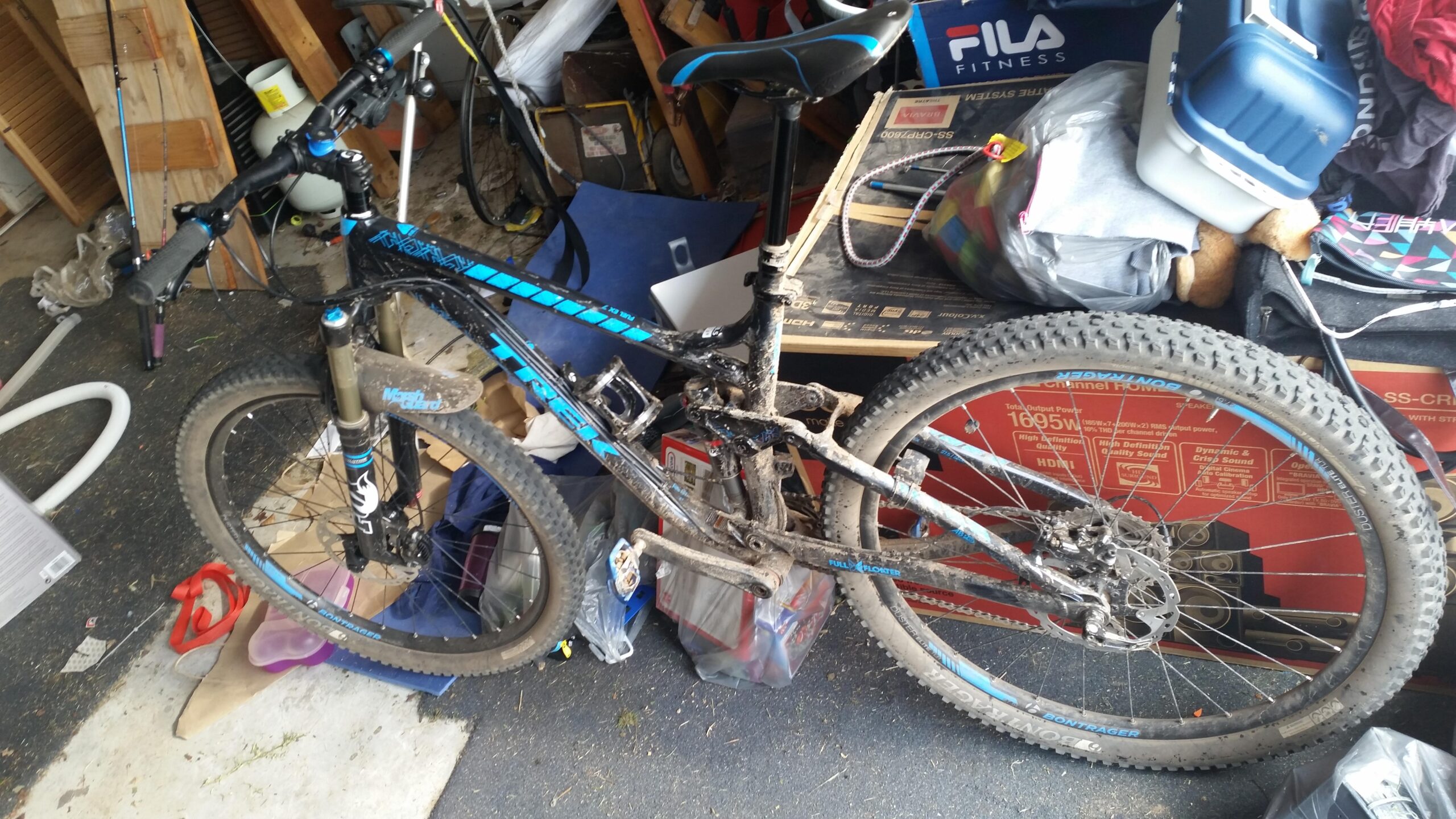 Trek Fuel EX 7: A mountain bike resting on a cluttered garage floor, featuring a blue and black frame, dirty tires, and worn components. Surrounding the bike are various items including boxes, a vacuum hose, and some clothing, highlighting the disorganized space.