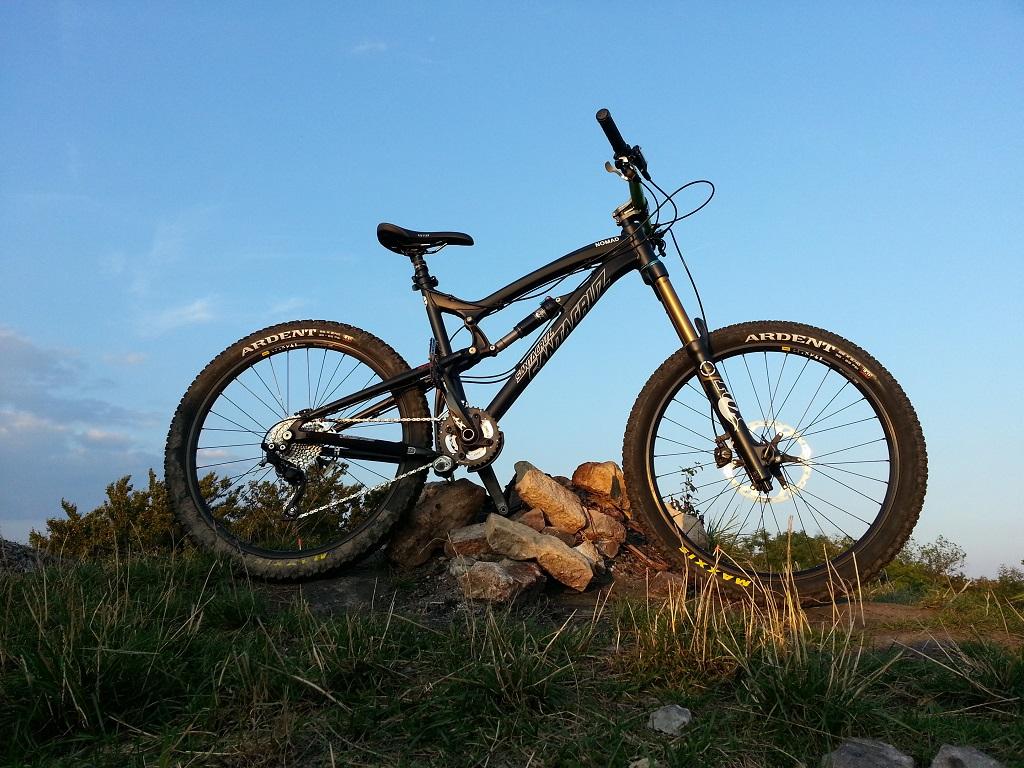 Santa Cruz Nomad: A mountain bike resting on a pile of rocks, set against a clear blue sky. The bike features a black frame and wide tires with "Ardent" branding. Grass and small shrubs are visible in the background, indicating an outdoor setting suitable for biking.