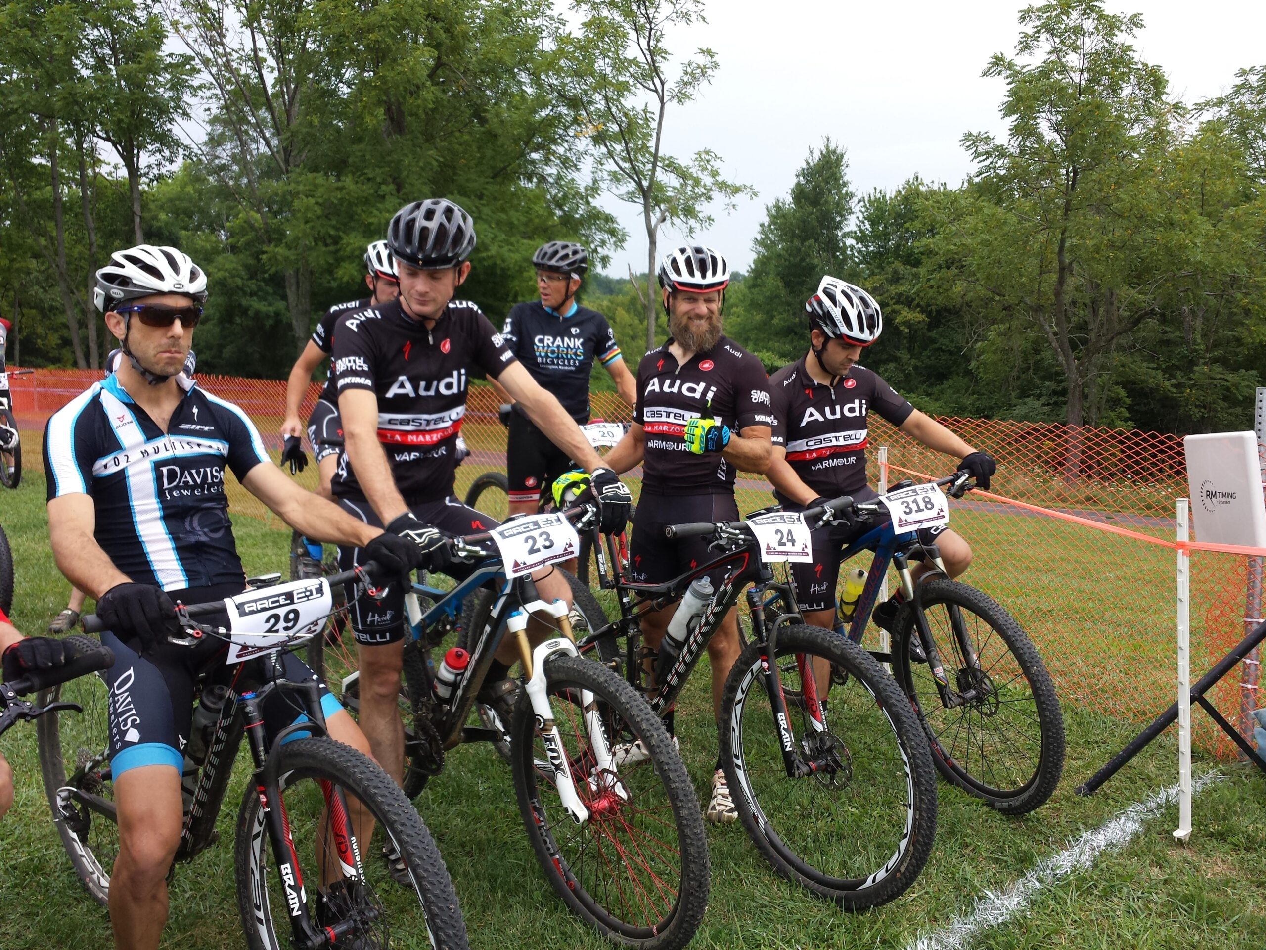 A group of five male mountain bikers, wearing competitive cycling jerseys and helmets, are lined up with their bikes before a race. Each cyclist has a race number displayed on their bike. The background features a grass field and trees, with an orange safety barrier visible. The sky is overcast, indicating potentially humid or rainy weather.