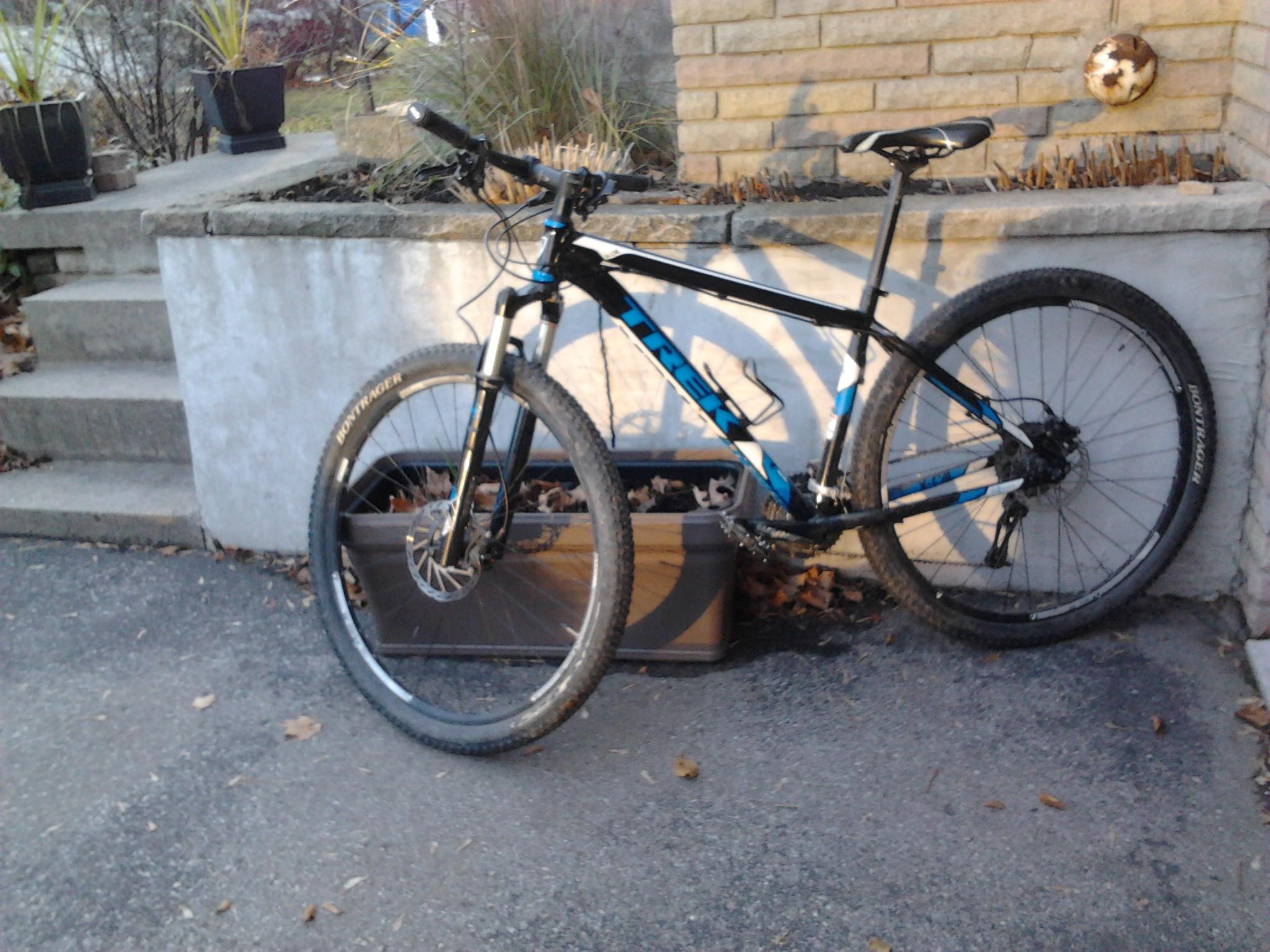 Trek X-Caliber: A black and blue mountain bike leaning against a concrete wall, with a planter box in the foreground and a few steps leading up to a garden area in the background. Fallen leaves are scattered on the ground.