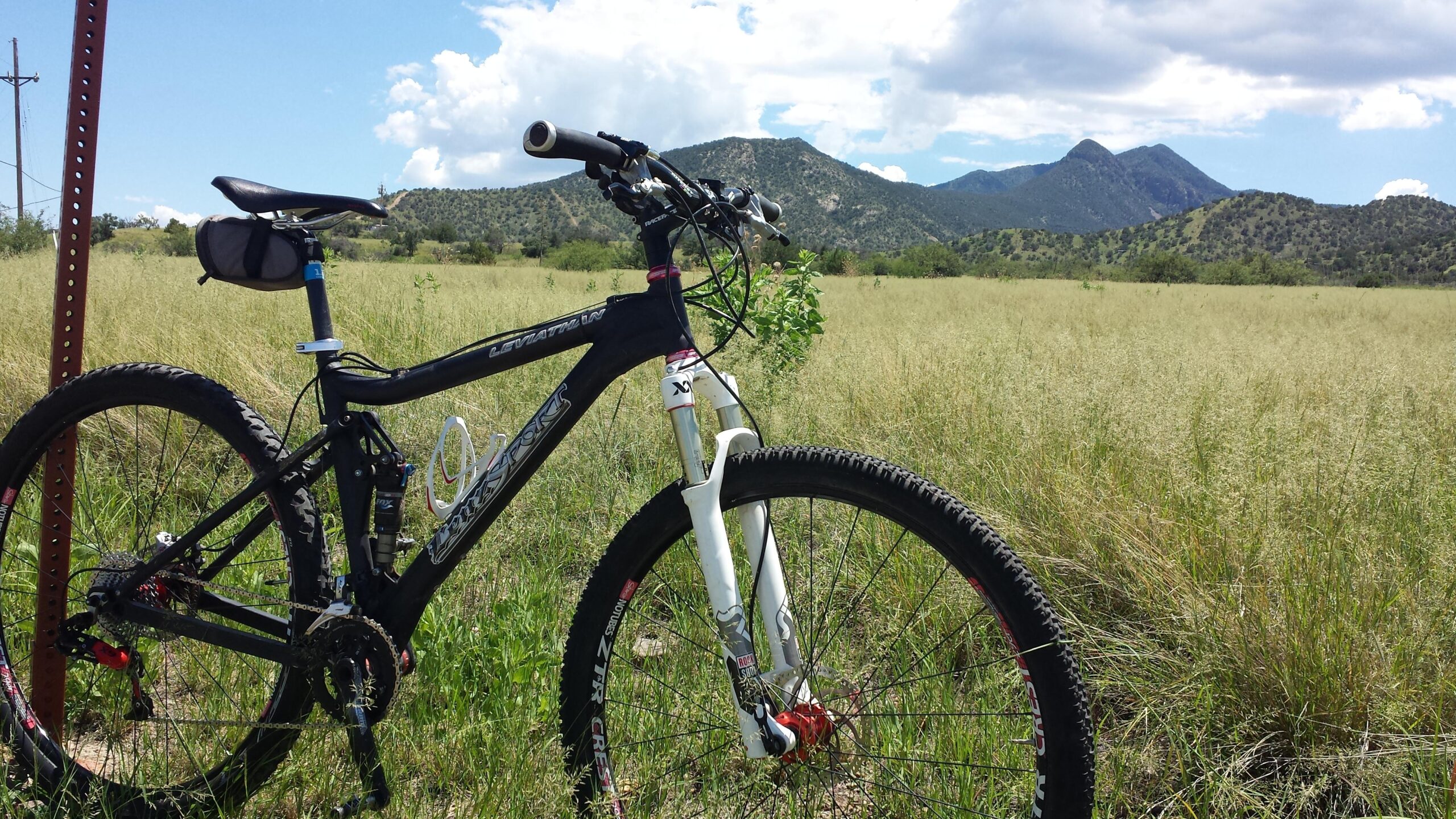 Lenz Sport Leviathan 4.0: A mountain bike resting on uneven terrain, with lush green grass surrounding it. In the background, rolling hills and distant mountains are visible under a partly cloudy sky. A signpost stands nearby, indicating the outdoor setting.