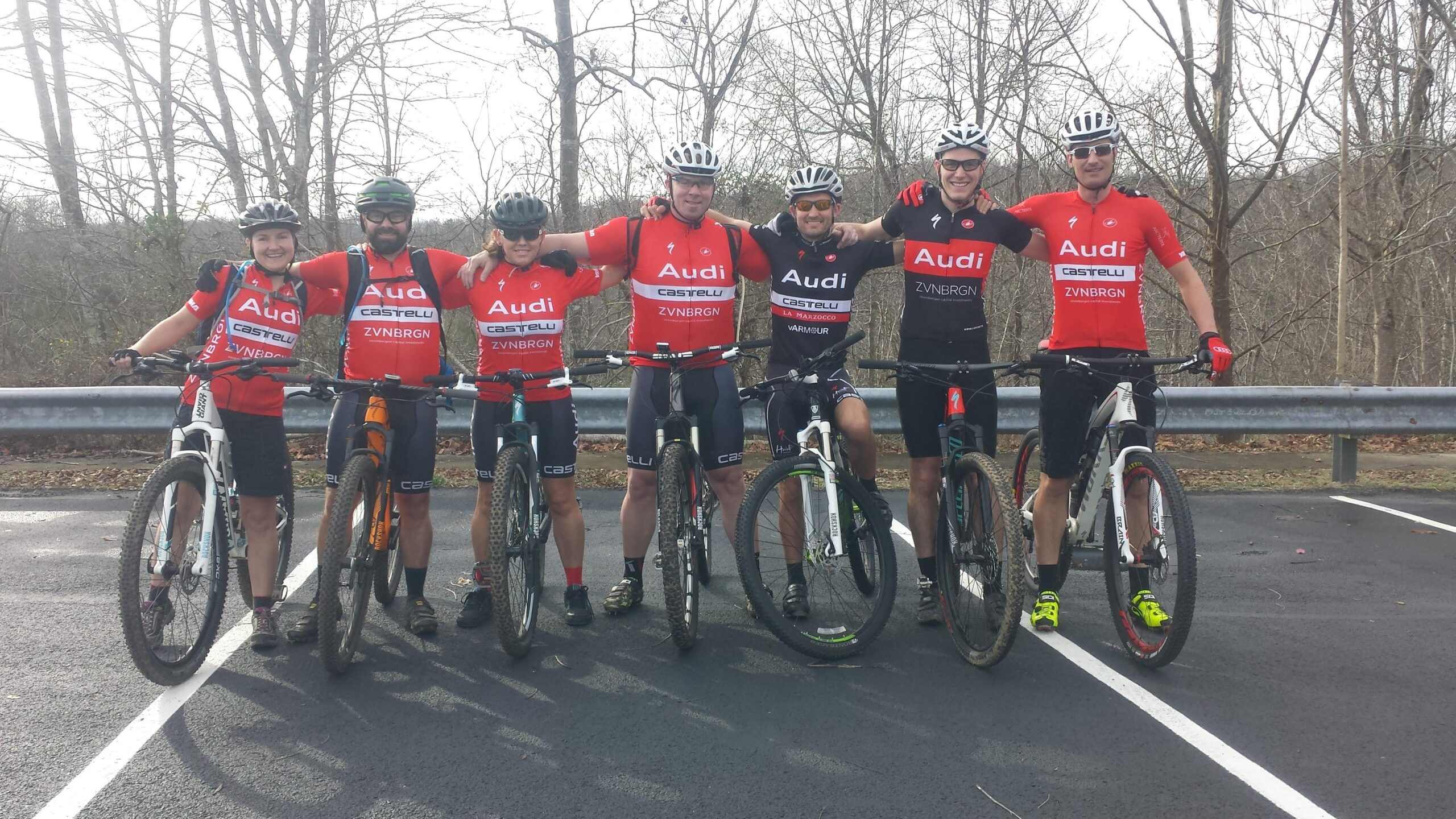 A group of seven mountain bikers in matching red and black cycling jerseys stand together, smiling and posing with their bikes. They are gathered near a roadside with sparse trees in the background, indicating a recreational outdoor environment. The cyclists appear to be excited and in a friendly mood after a ride.