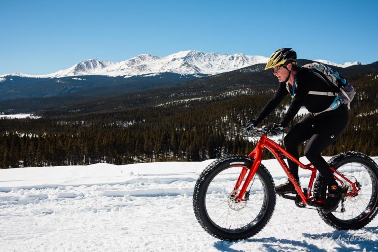 A person riding a fat tire bicycle through a snowy landscape with distant mountains and a clear blue sky in the background. The cyclist is wearing a helmet and black clothing, with a backpack, and is navigating the snow-covered terrain.
