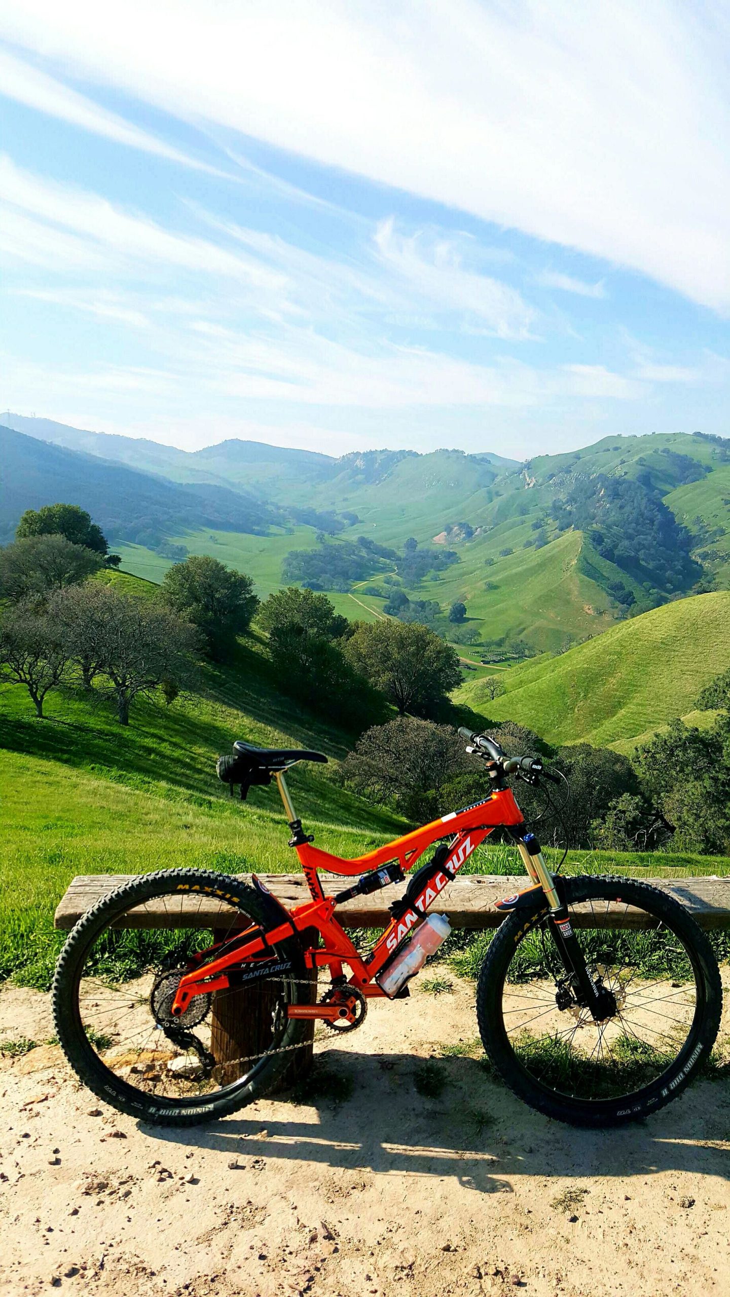 A bright orange mountain bike rests on a wooden bench, overlooking lush green hills and rolling landscapes under a blue sky with wispy clouds. Black Diamond Mines mountain bike trail.