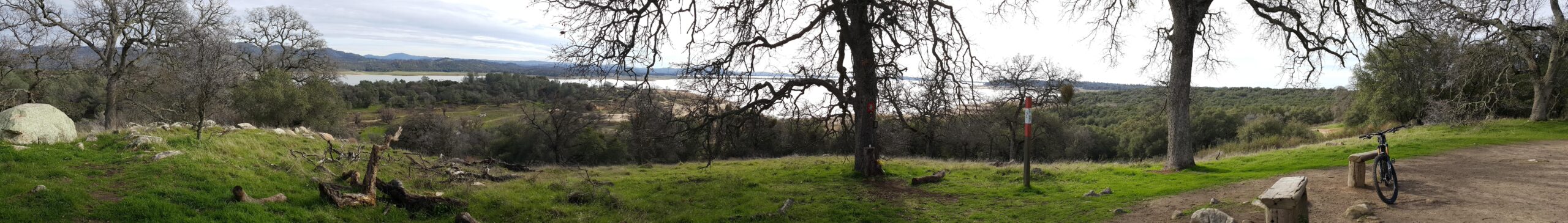 A panoramic view of a natural landscape featuring rolling hills, a lake in the distance, and scattered trees. In the foreground, there is a grassy area with a dirt path, a wooden bench, and a mountain bike resting nearby. The sky is overcast, lending a soft light to the scene, while the lush greenery contrasts with the earthy tones of the ground. Granite Bay Trail mountain bike trail.