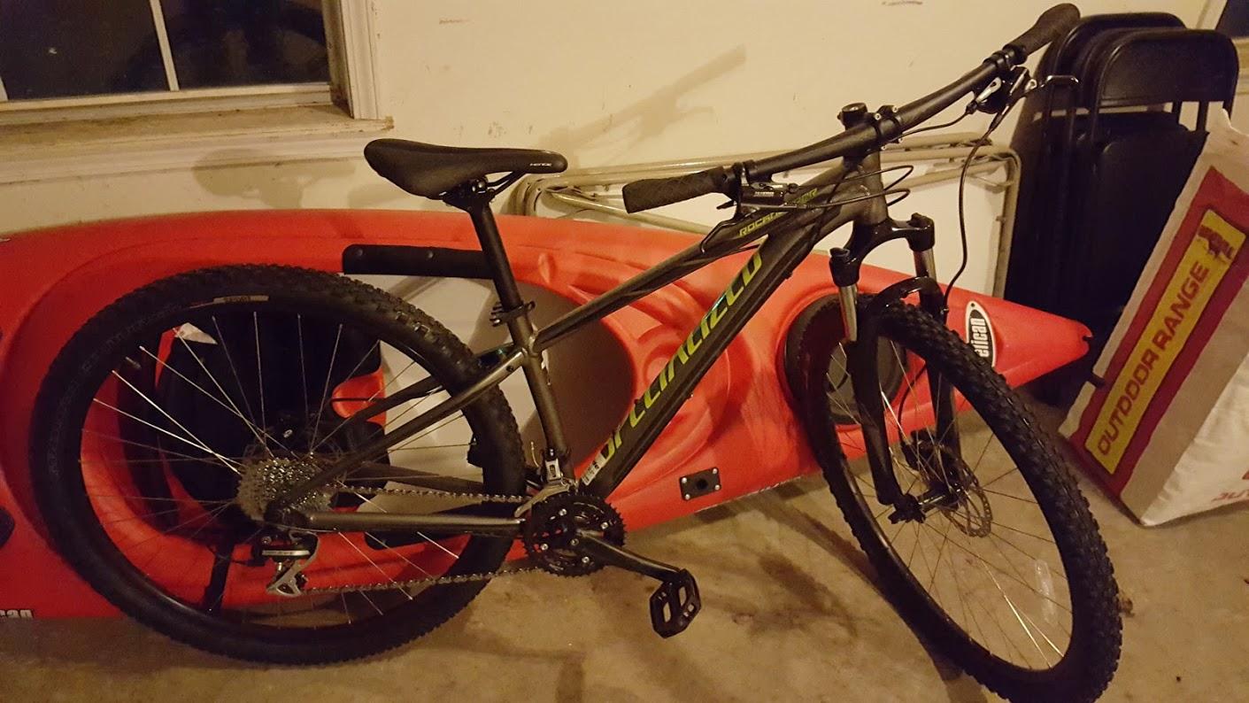 Specialized Rockhopper 29: A mountain bike in a garage setting, positioned next to a red kayak and various other storage items. The bike features a black and gray frame with green accents, sturdy tires, and a visible gear system.