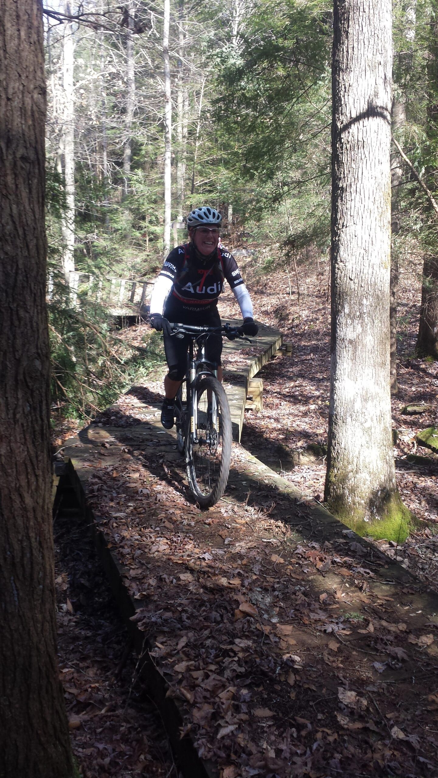 A cyclist rides a mountain bike on a narrow wooden bridge surrounded by trees in a forested area. The ground is covered in fallen leaves, and the cyclist is wearing a helmet and biking gear. Sunlight filters through the trees, highlighting the trail ahead.