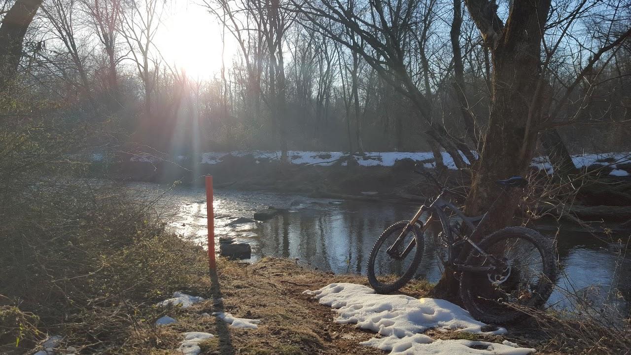 A mountain bike leans against a tree along a riverbank, with the sun shining low in the sky. Snow is scattered on the ground, and the surrounding trees are bare, indicating early spring or late winter. The water reflects the sunlight, creating a serene nature scene. Seneca Bluffs Trail mountain bike trail.