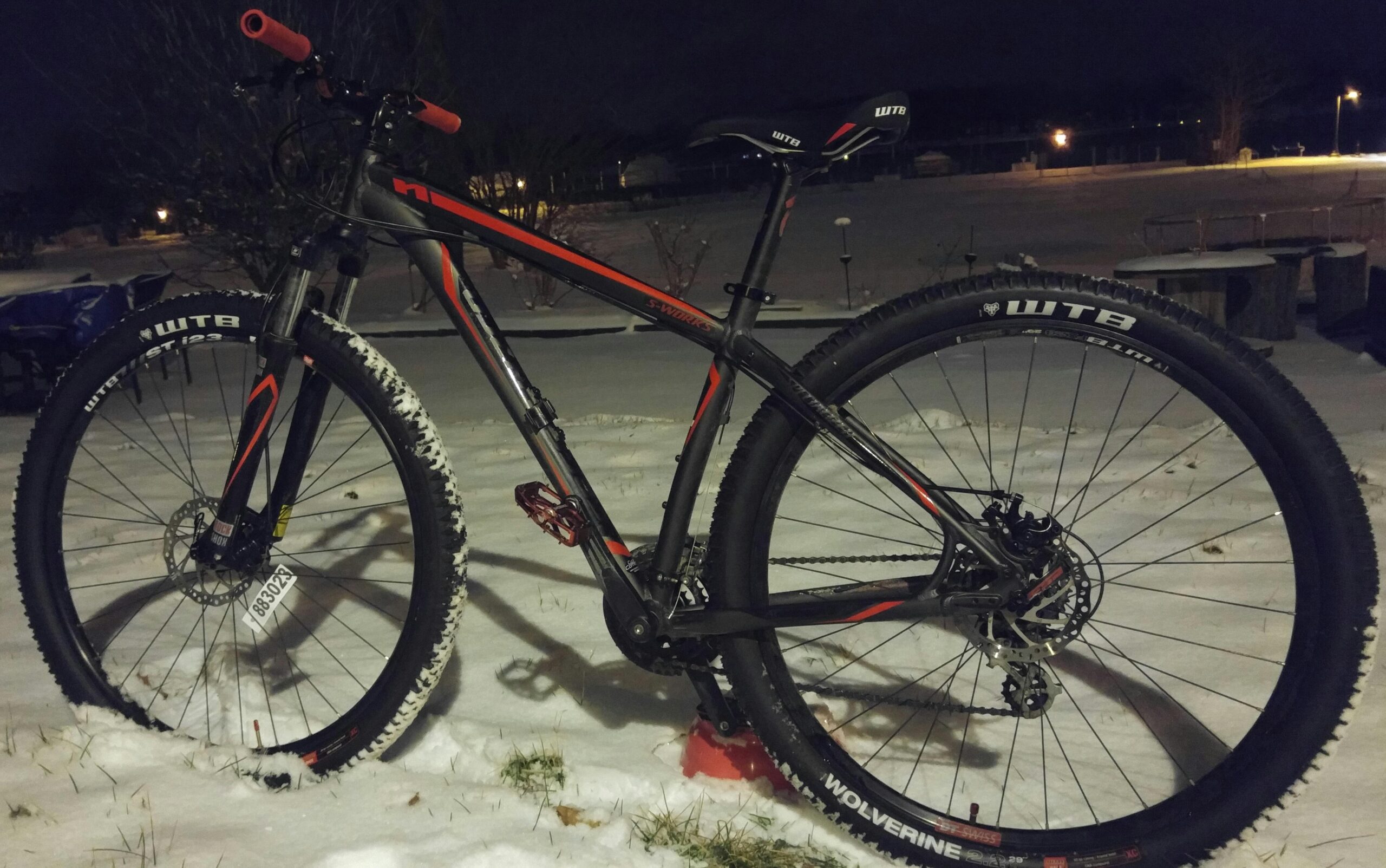 Specialized Hardrock Sport Disc 29er: A mountain bike with a sleek black and orange frame is positioned on a snowy landscape at night. The bike features chunky tires with visible tread, partially covered in snow, and is equipped with disc brakes. In the background, soft lights illuminate the scene, showcasing a tranquil winter setting.