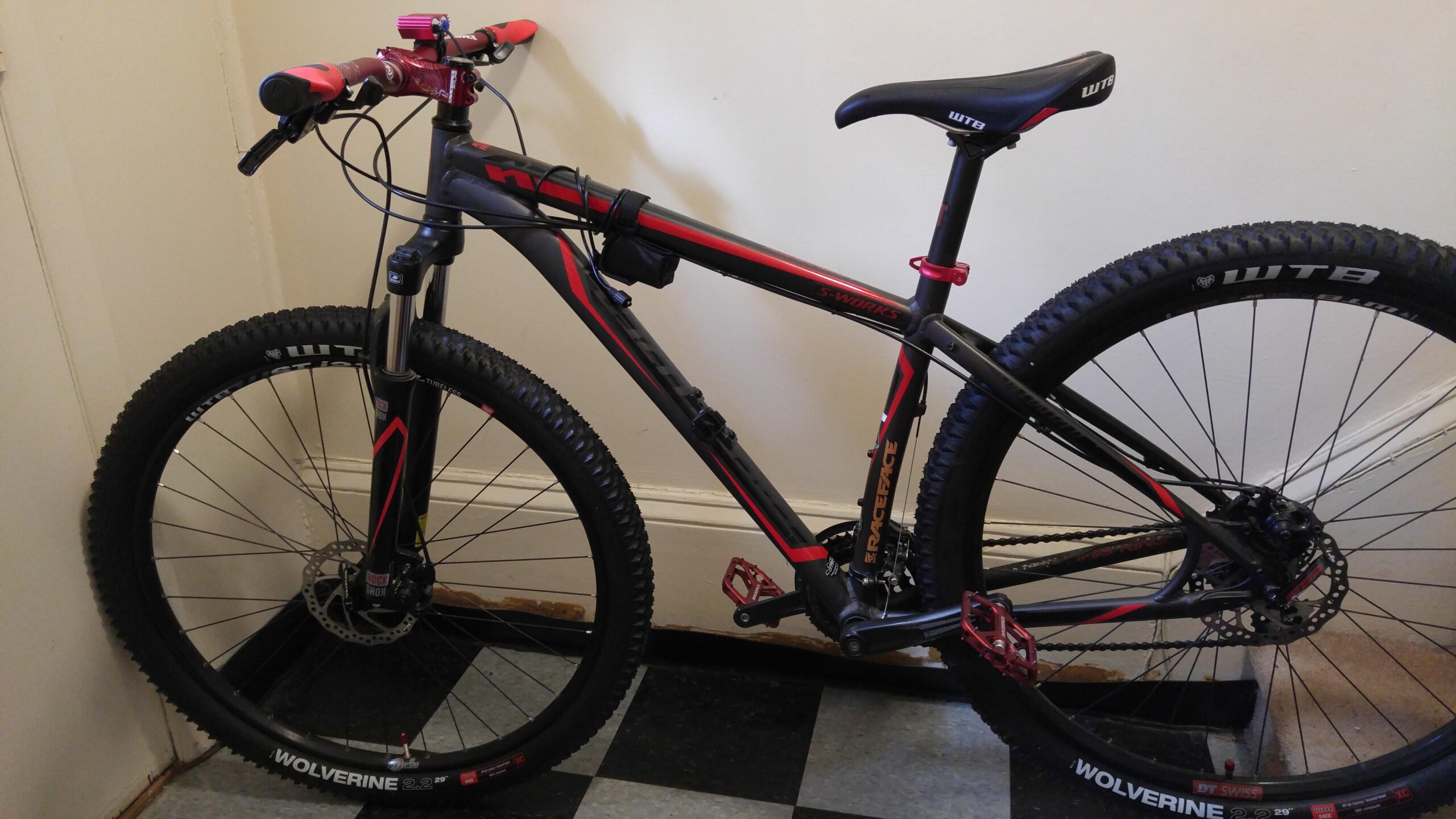 A black and red mountain bike leaning against a wall, showcasing its features such as thick tires, a suspension fork, and a sleek frame design. The handlebars have red grips, and the pedals are also highlighted in red. The background consists of a simple indoor setting with a checkered floor.