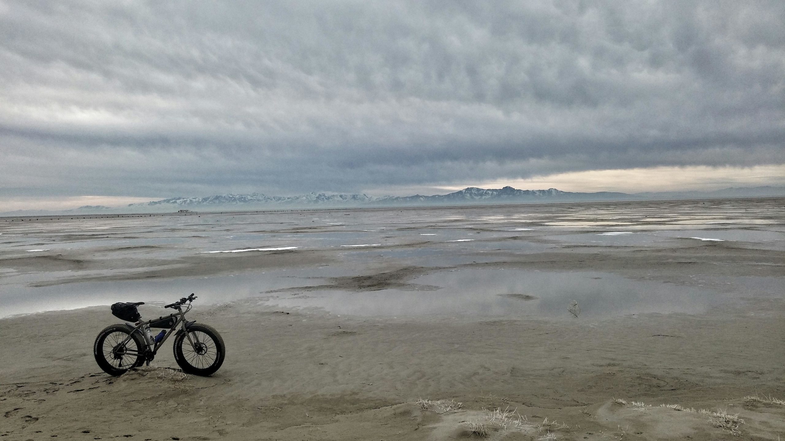 A fat-tire bike is parked on a sandy shore, with a vast expanse of wetland and reflections in shallow water. In the background, snow-capped mountains are visible under a cloudy sky. The scene conveys a serene and remote outdoor environment. Antelope Island mountain bike trail.