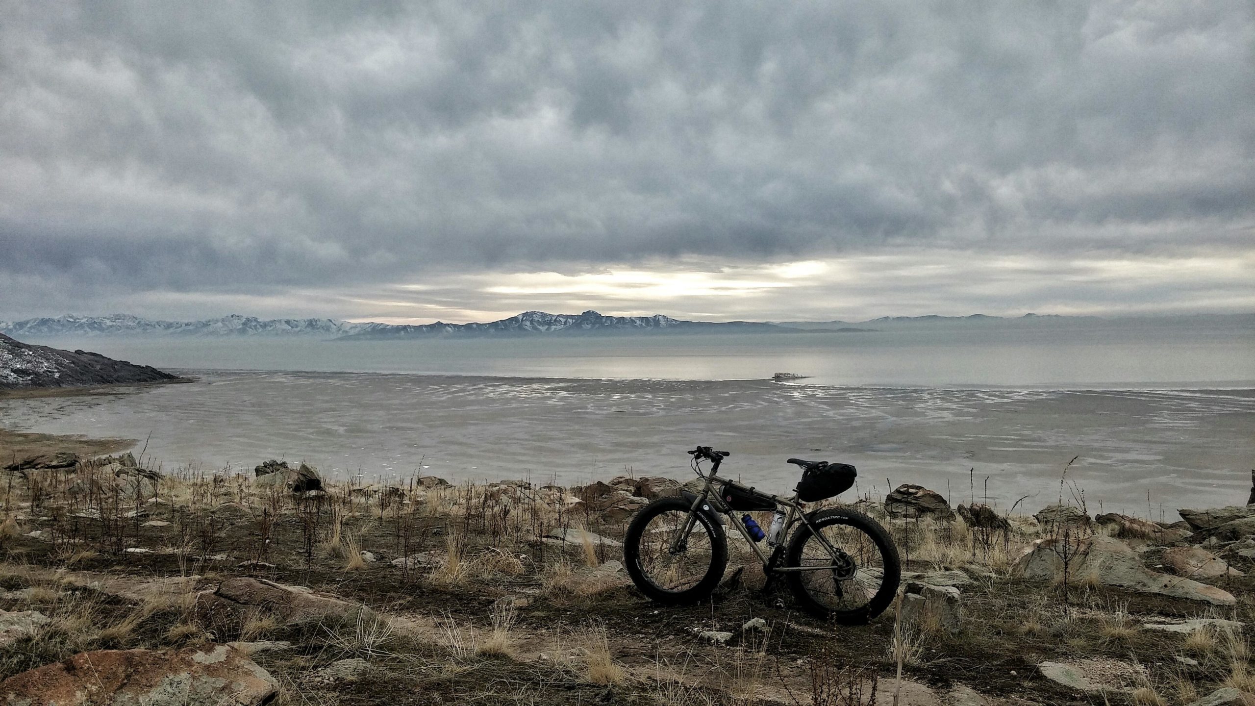 A fat bike stands on rocky terrain overlooking a lake, with distant mountains visible under a cloudy sky. The landscape is characterized by a mix of dry grass and sparse vegetation, with the water reflecting a muted light. Antelope Island mountain bike trail.