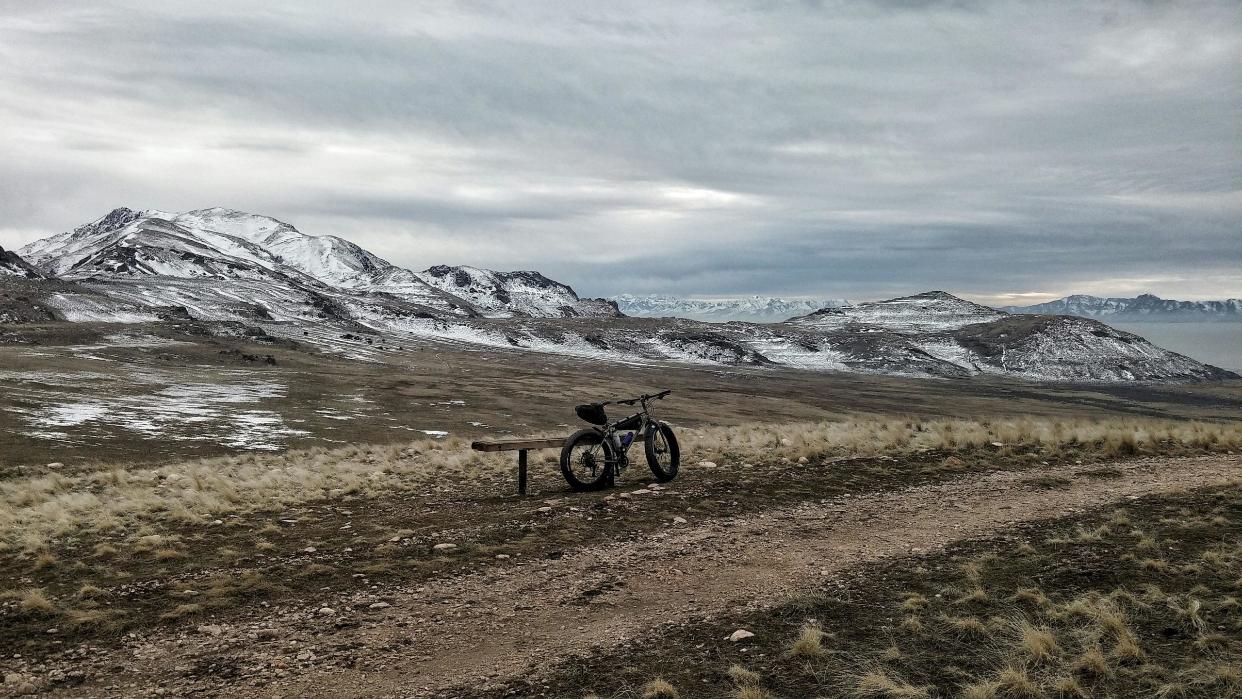 A mountain bike leaning against a wooden bench on a rocky trail, surrounded by vast grassland and snow-capped mountains under a cloudy sky. Antelope Island mountain bike trail.