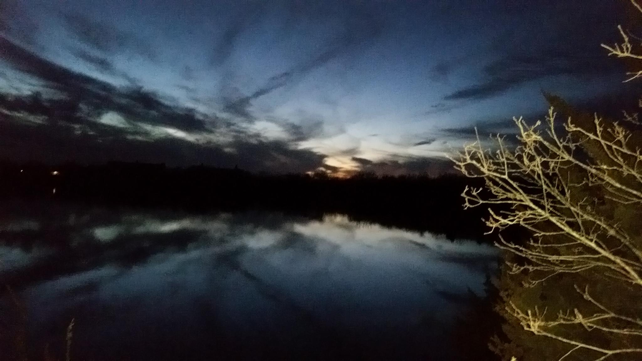 A tranquil evening scene depicting a calm body of water reflecting the twilight sky, with scattered clouds and a hint of light from the setting sun on the horizon. In the foreground, a bare tree branch is visible, adding to the serene atmosphere of the dusk setting. Lehigh Portland Trails mountain bike trail.