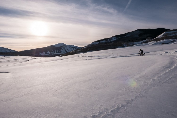 A lone cyclist rides a fat bike across a snow-covered landscape under a bright winter sun, with snow-capped mountains in the background and a clear sky. The scene captures the serene beauty of winter outdoor activities.