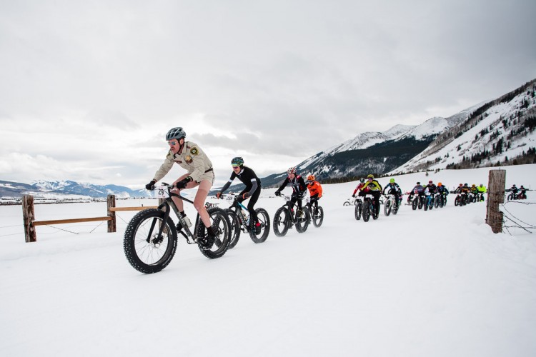 A group of cyclists participate in a fat bike race on a snowy landscape, with mountains in the background. The lead cyclist wears a unique costume while the others are dressed in various athletic gear, showcasing the fun and diverse atmosphere of the event.