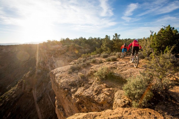 Two mountain bikers riding along a rocky trail on a cliff edge during sunset, surrounded by sparse vegetation and trees, with a scenic view of the canyon below.