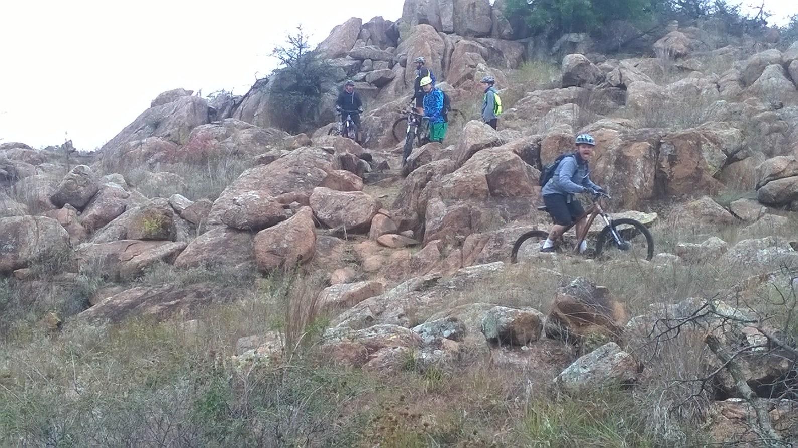 A group of five mountain bikers navigating a rocky and uneven terrain, surrounded by patches of grass and boulders. The riders are focused as they maneuver their bikes over the challenging landscape, with some climbing uphill among large rocks. The scene captures the thrill of outdoor cycling in a natural setting. Lake Lawtonka Trails mountain bike trail.