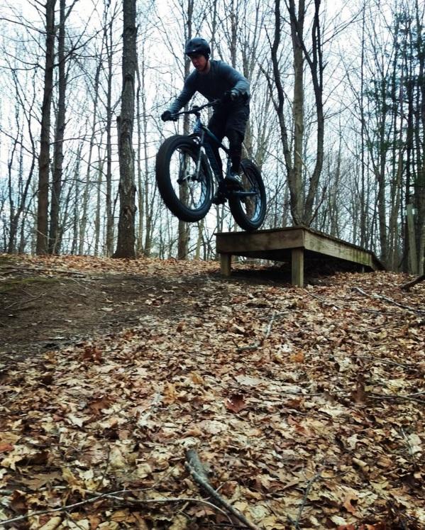 A person riding a fat bike jumps off a wooden platform in a wooded area during autumn, surrounded by fallen leaves. The rider is airborne, demonstrating a dynamic biking maneuver in a natural setting. Anderson Park mountain bike trail.