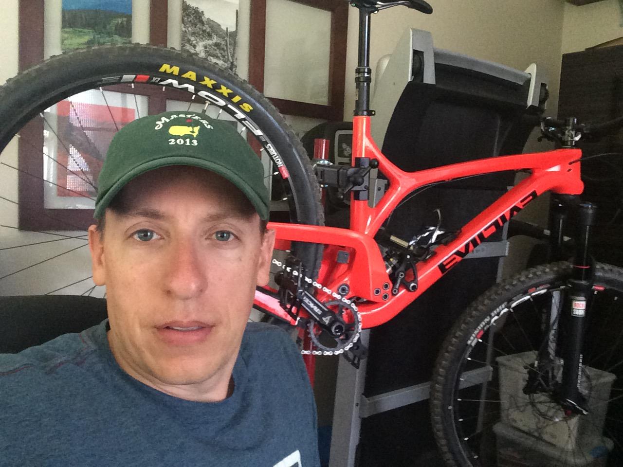Evil The Following: A person sitting in a chair is partially visible in the foreground, with a mountain bike in bright red behind them. The bike features thick tires and visible components. The background includes framed pictures and clutter typical of a home workspace. The individual wears a green cap and a blue shirt.