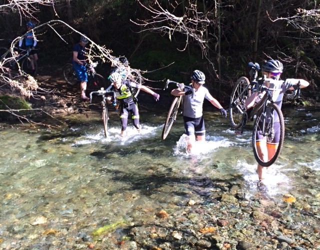 A group of cyclists wading through a shallow stream while carrying their bicycles. The scene is surrounded by trees, with sunlight filtering through the branches, highlighting the clear water and rocky bed. Several cyclists are visible in the background, continuing their journey.
