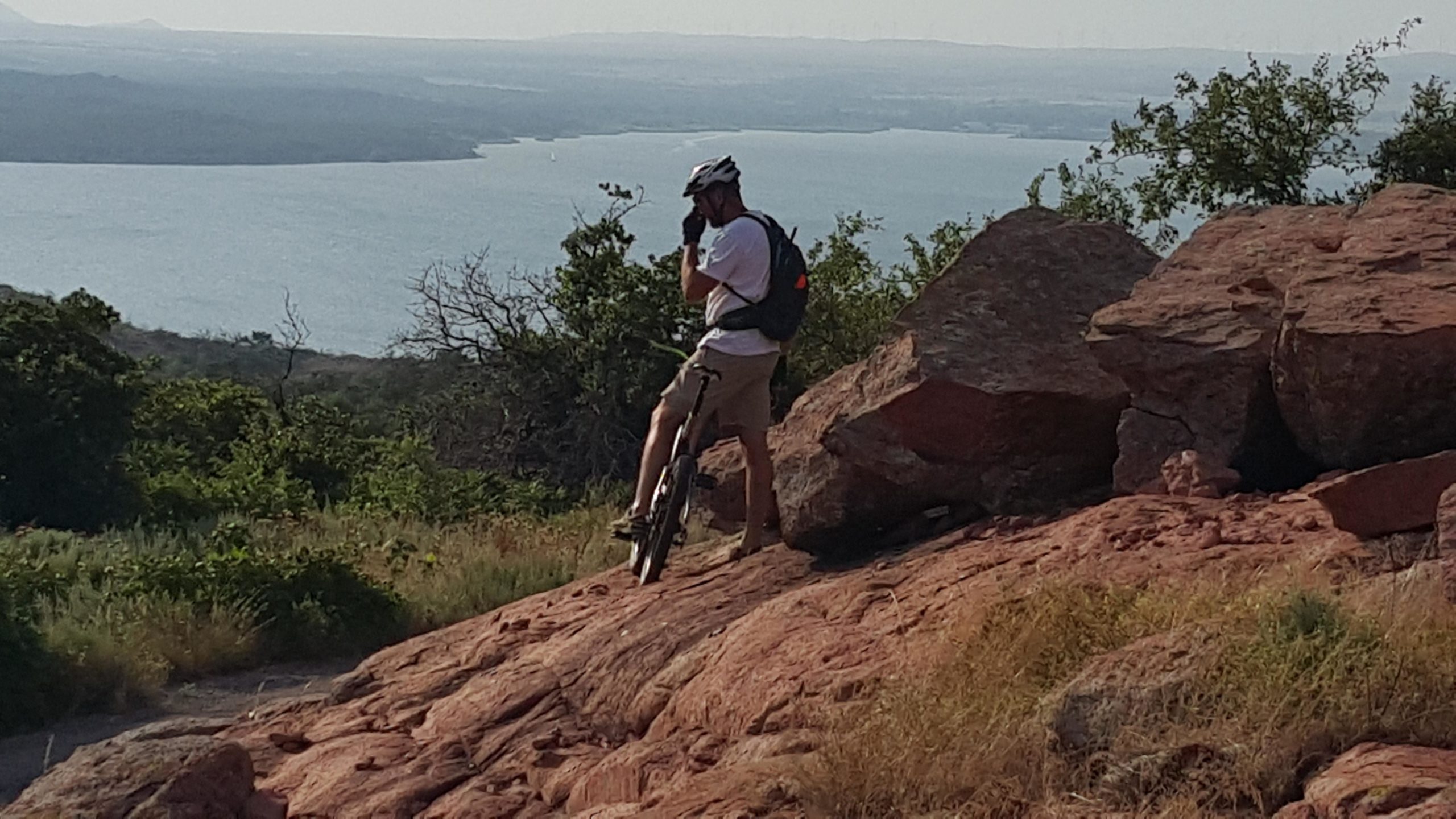 A person wearing a helmet and backpack stands beside a mountain bike on rocky terrain, overlooking a body of water and distant hills under clear skies. Lake Lawtonka Trails mountain bike trail.