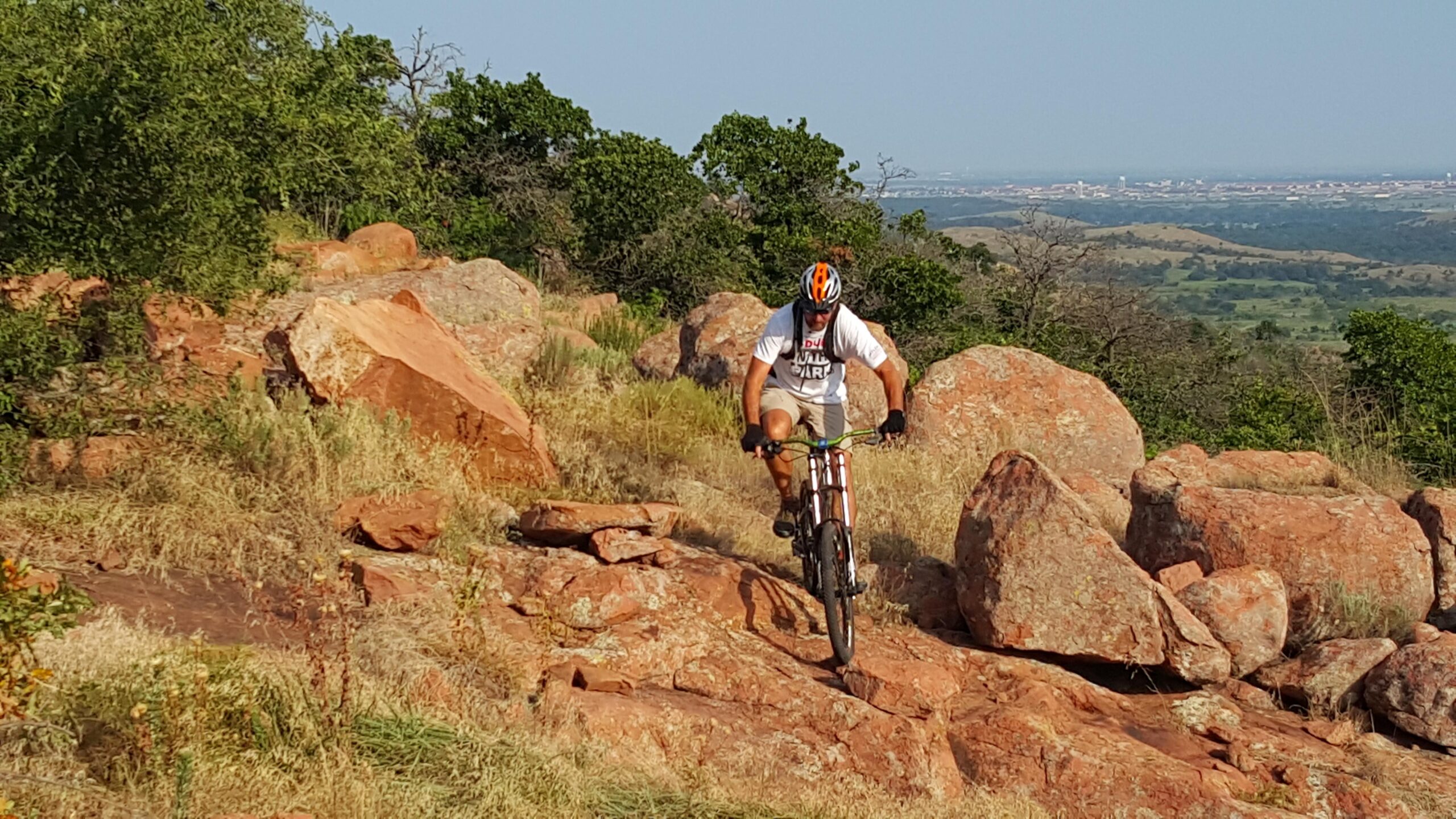 A mountain biker navigating rocky terrain on a sunny day, surrounded by green foliage and distant hills. The biker is wearing a helmet and a t-shirt while carefully maneuvering over large red rocks. Lake Lawtonka Trails mountain bike trail.