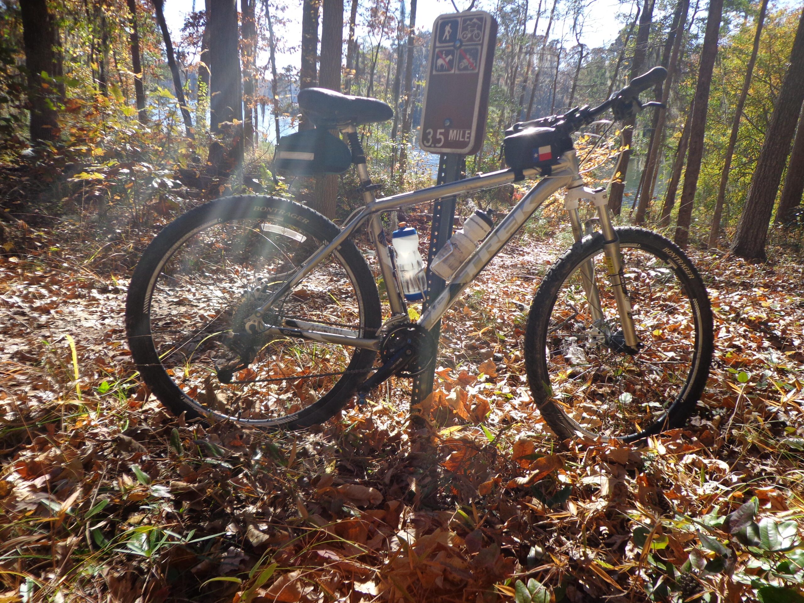 Trek Marlin: A silver mountain bike resting on a leafy forest floor, with a trail sign indicating a 35-mile bike route in the background. Sunlight filters through the trees, illuminating the scene, which features autumn foliage. The bike has a water bottle attached to it and a small bag on the seat.