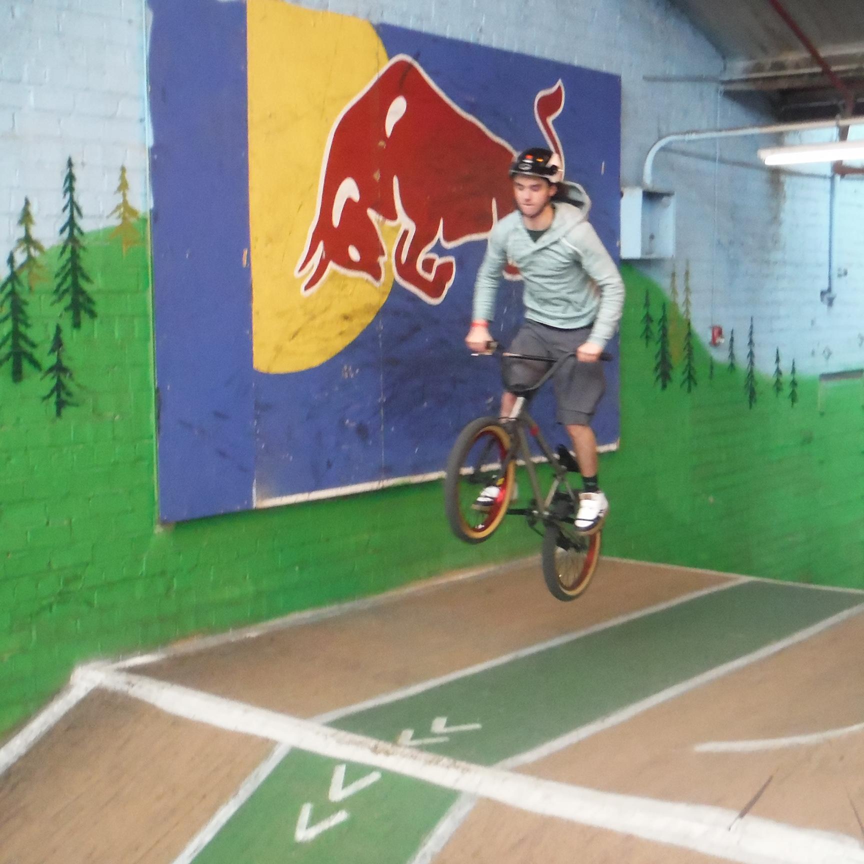 A person performing a BMX trick in a skatepark, soaring over a ramp with a colorful Red Bull logo painted on the wall behind them. The scene features green painted walls adorned with trees and a wooden ramp with marked lanes. Rays Indoor Mtb Park mountain bike trail.