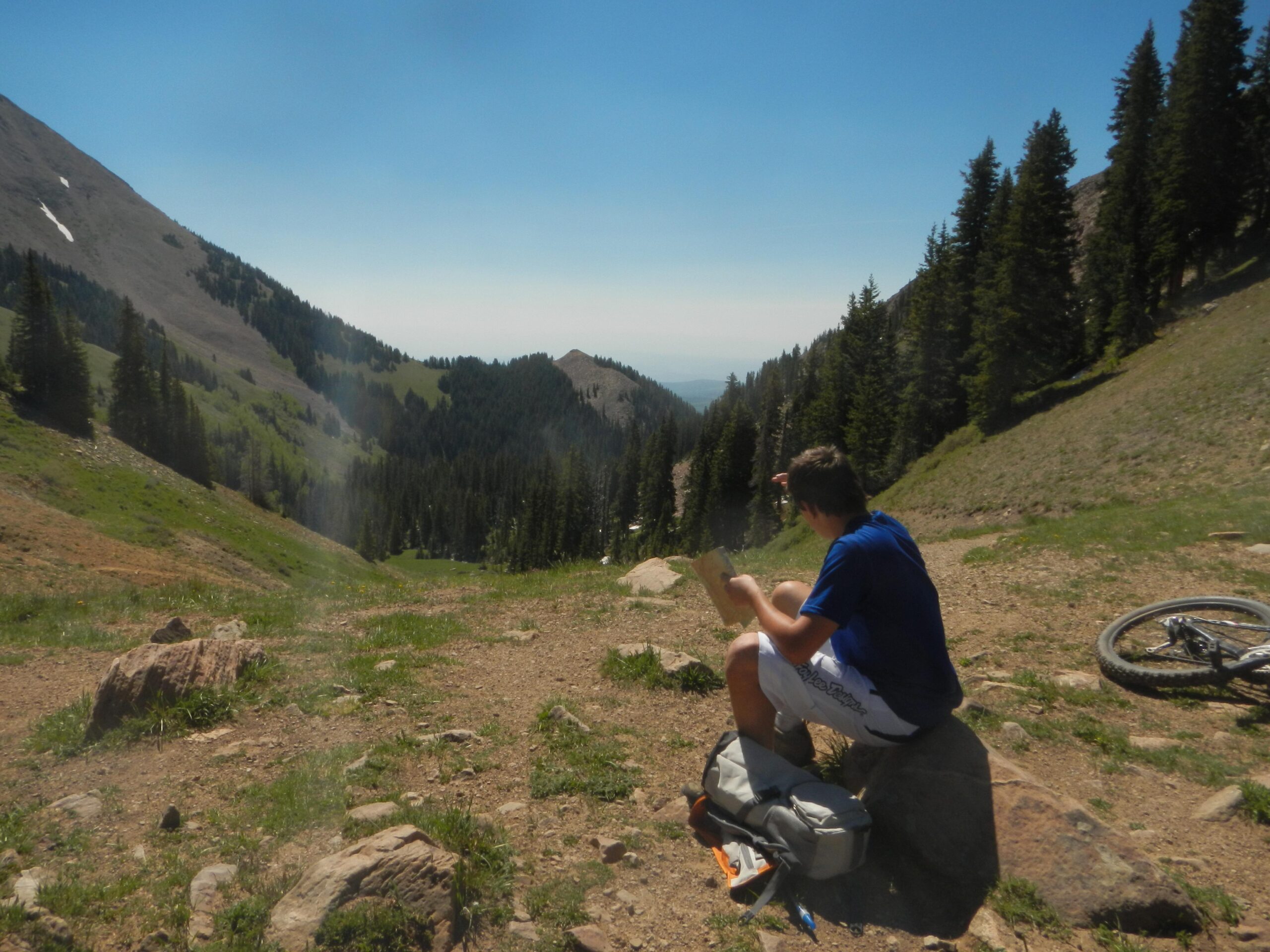 A person sitting on a rock in a mountainous area, wearing a blue shirt and shorts, is observing a landscape filled with green hills and tall trees. A mountain bike is leaning against a nearby rock, and the sky is clear and blue, indicating a sunny day. The Whole Enchilada mountain bike trail.