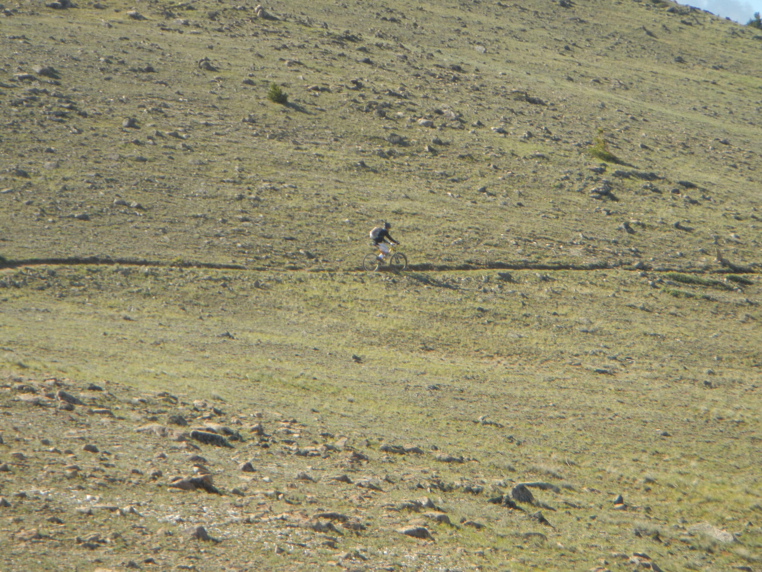 A mountain biker riding along a narrow trail on a rocky hillside, surrounded by sparse vegetation and open landscape. Monarch Crest Trail mountain bike trail.