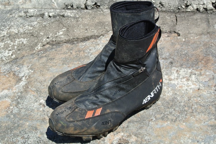 A pair of black and orange cycling boots with a zipper on the side, resting on a textured rock surface. The boots appear worn, with some dirt and scuff marks visible.
