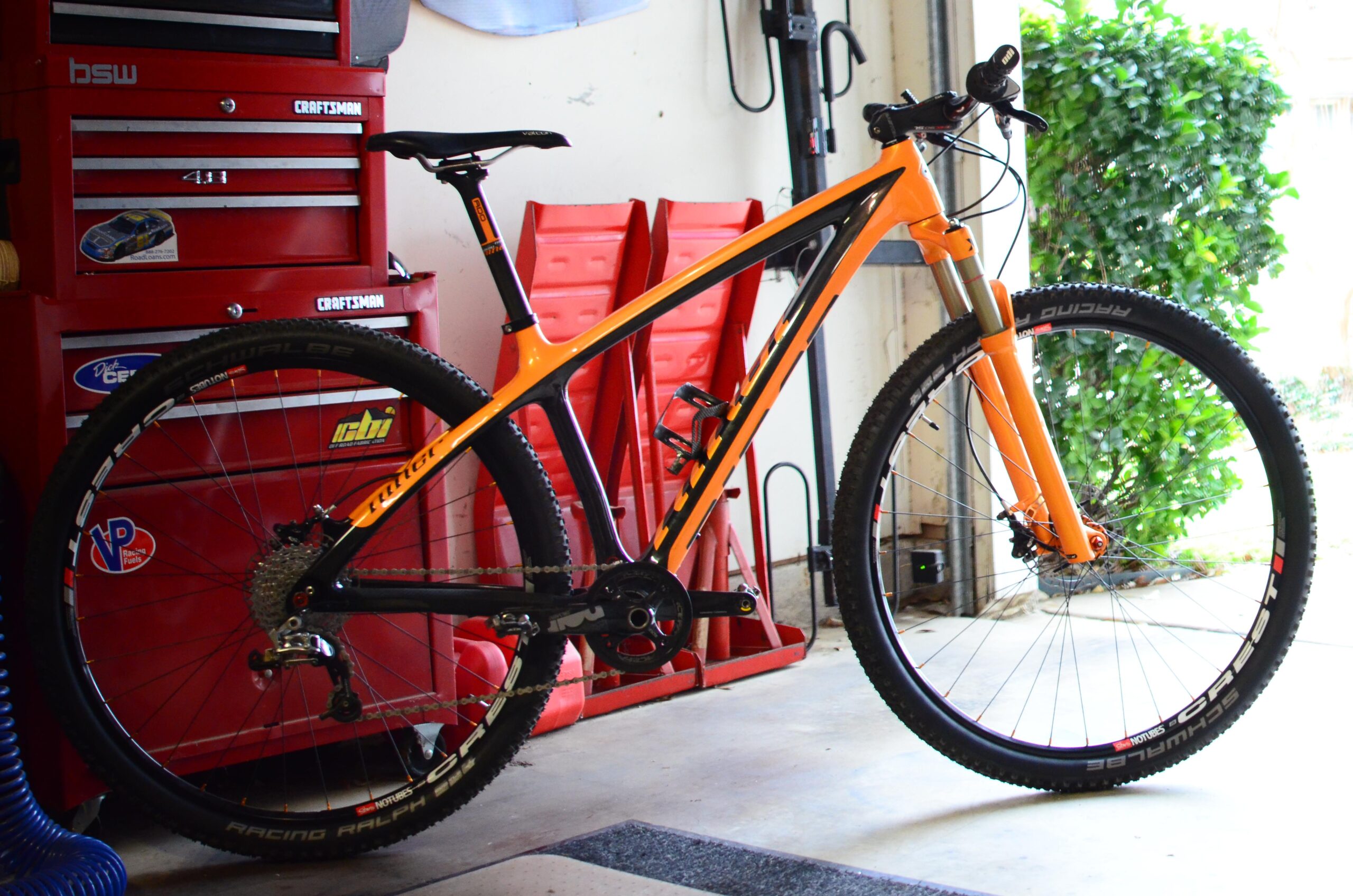 Niner Air 9: A bright orange mountain bike with black accents parked in a garage. The bike features a lightweight frame, thick tires for off-road riding, and a visible chain and derailleur. In the background, there are red tool cabinets and greenery visible outside the garage.