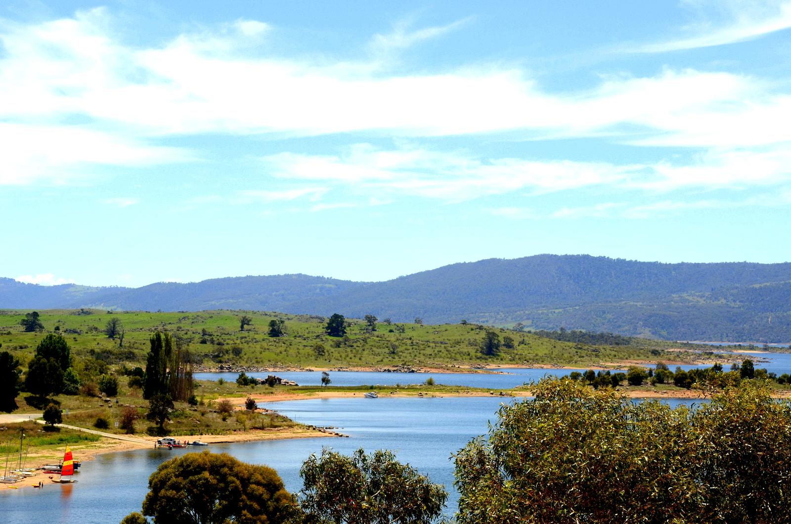 A serene landscape featuring a calm lake surrounded by lush greenery and rolling hills. The sky is clear with a few clouds, and there are small boats visible on the water near a sandy shore. Tall trees and shrubs frame the foreground, creating a picturesque view of nature. Jindabyne Community Trail mountain bike trail.