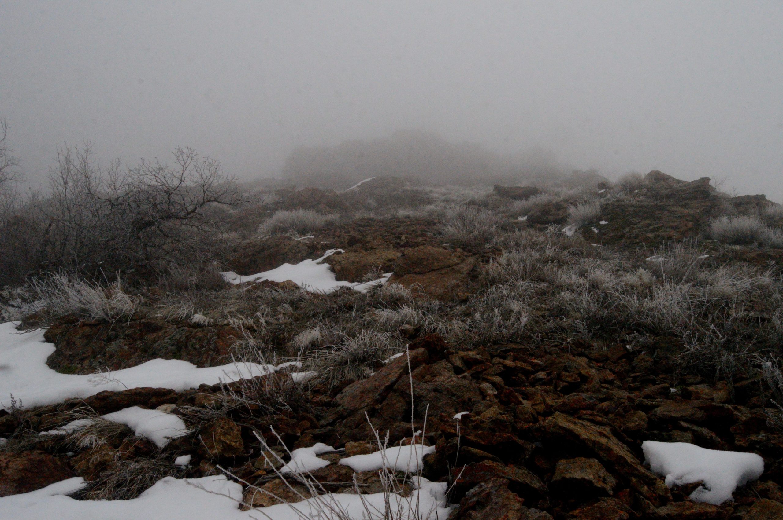 A rocky landscape shrouded in dense fog, with patches of snow and sparse vegetation. The view is obscured at a distance, creating a mysterious atmosphere. Bonneville Shoreline Trail - Ogden Section mountain bike trail.
