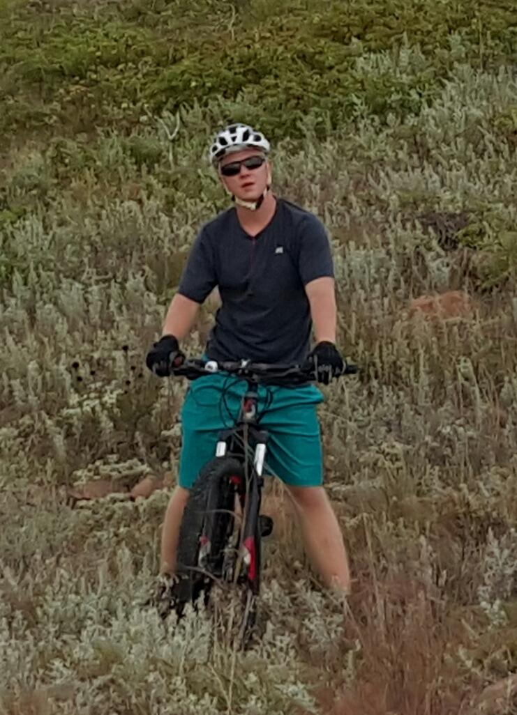 A person wearing a helmet and sunglasses is standing on a mountain bike in a grassy area, looking toward the camera. They are dressed in a dark shirt and teal shorts, with gloves on their hands. The background features lush greenery and rocky terrain. Lake Lawtonka Trails mountain bike trail.