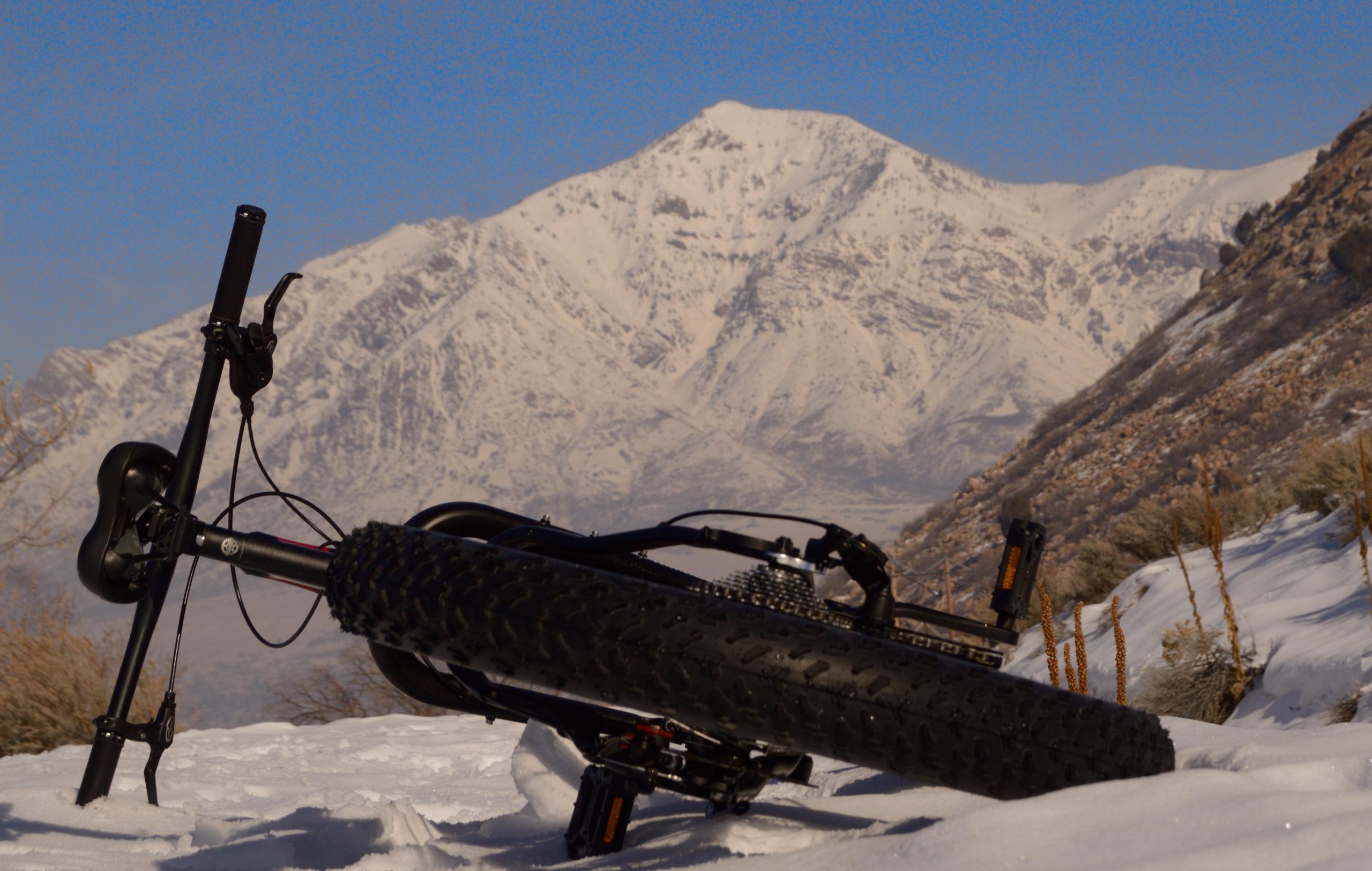 A fat bike lies on its side in a snowy landscape, with snow-covered mountains in the background under a clear blue sky. Bonneville Shoreline Trail - Ogden Section mountain bike trail.