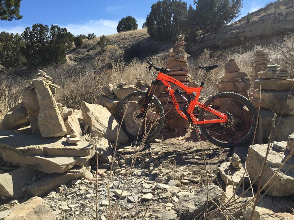 Foes Mixer Enduro: A bright orange mountain bike is positioned among rock formations on a dirt path, surrounded by sparse vegetation and trees in a sunny outdoor setting.