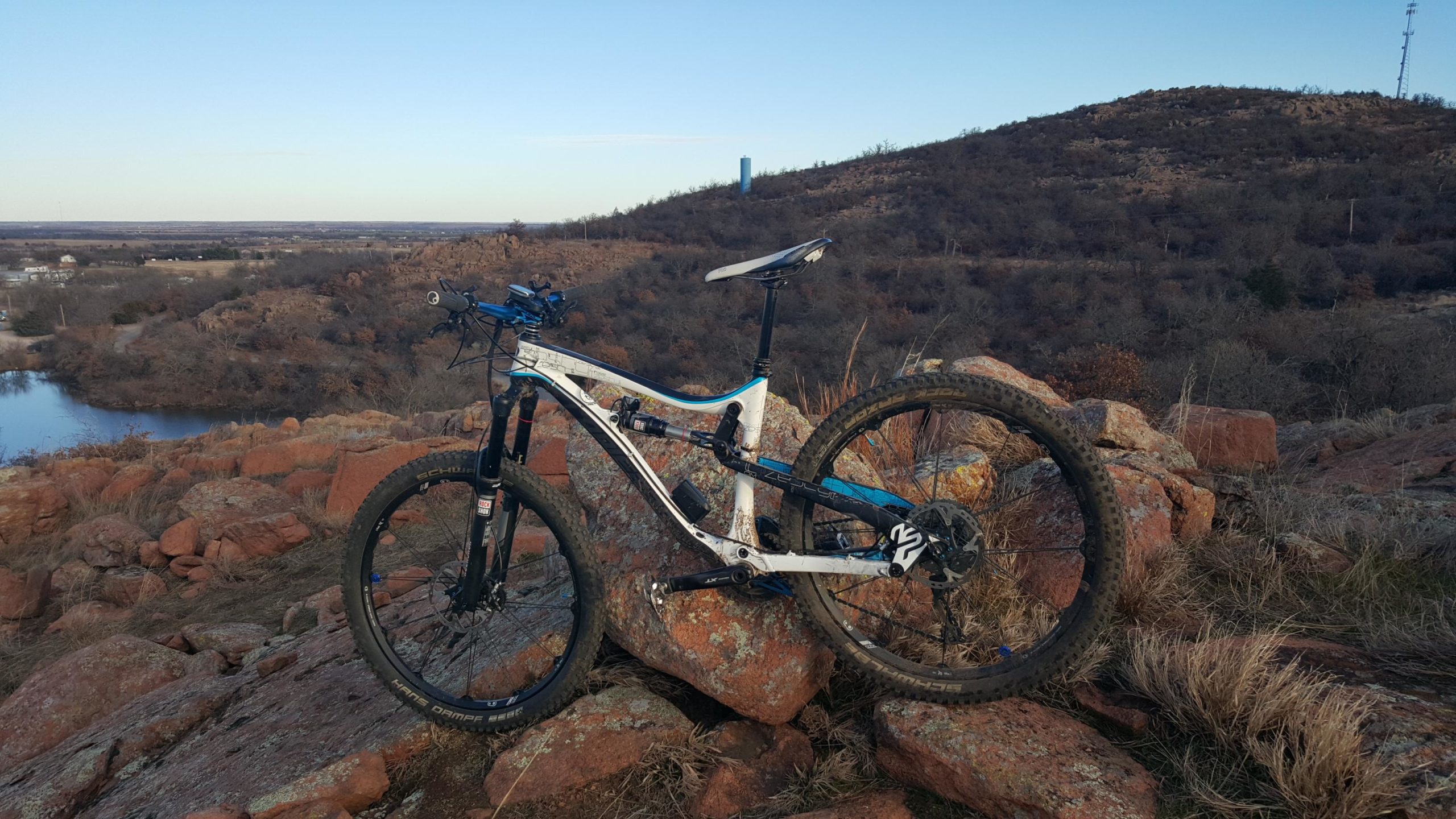 A mountain bike resting on a rocky outcrop with a picturesque view of the landscape, including a small body of water and distant hills under a clear sky during sunset. Lake Lawtonka Trails mountain bike trail.