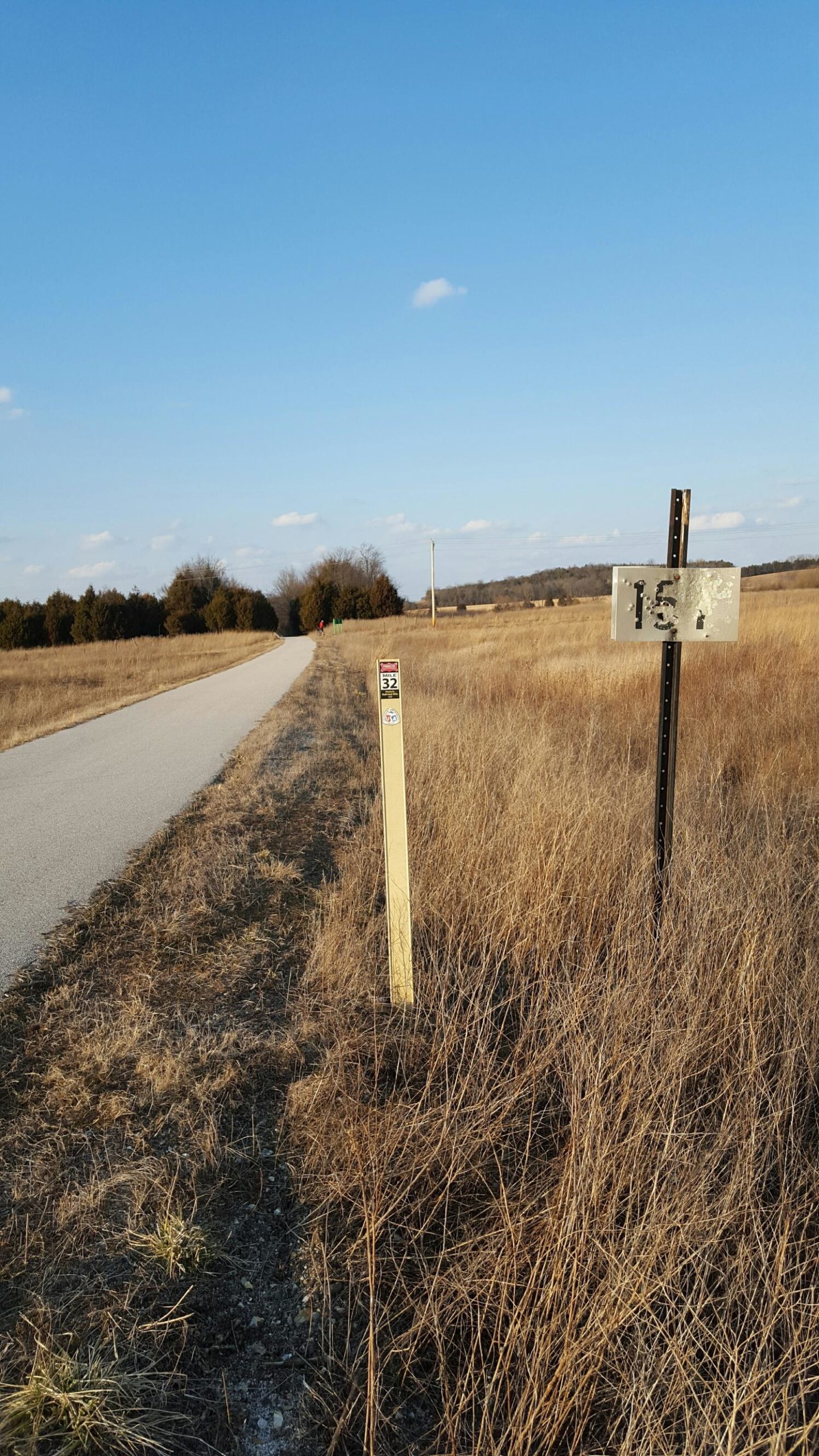 A dirt path stretches through an open, grassy landscape under a clear blue sky. On the right, two signposts are visible: one labeled "15A" with some wear and tear, and another with a small sticker, labeled "32." The area appears rural, with sparse trees in the background and a distant hint of utility poles. Frisco Highline Trail mountain bike trail.