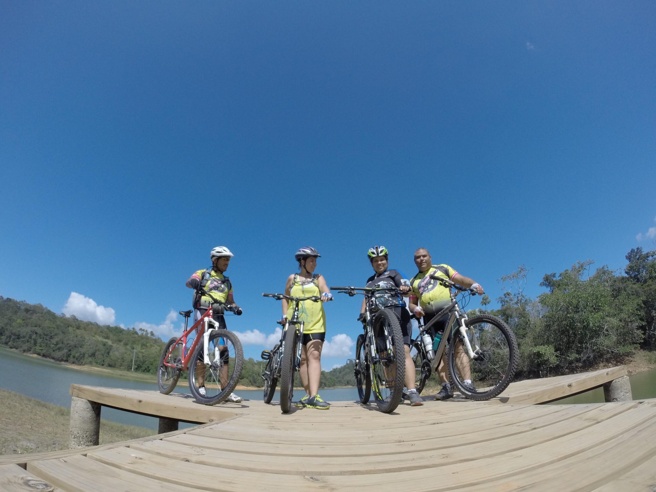 Specialized Hardrock 26: A group of four cyclists wearing bright yellow shirts and helmets, standing on a wooden dock by a serene body of water. The sky is clear and blue, with a few clouds visible in the background. The group is posed with their bicycles, enjoying a day of outdoor biking.